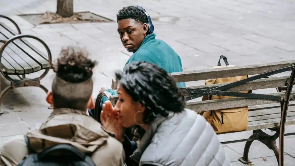 Two people whisper on a park bench while a man in the background looks back.