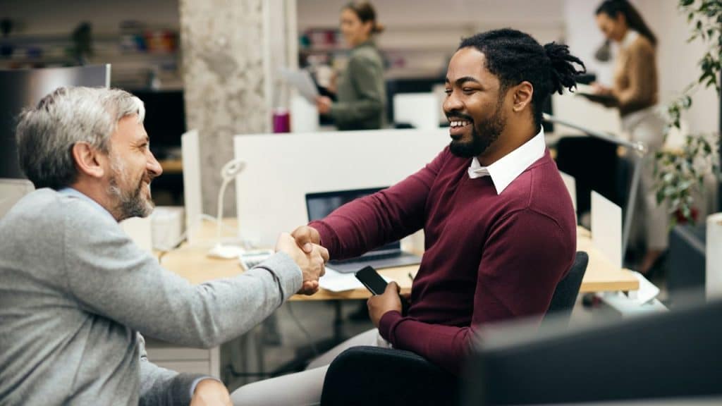 Two men in an office smile and shake hands while seated at their desks.
