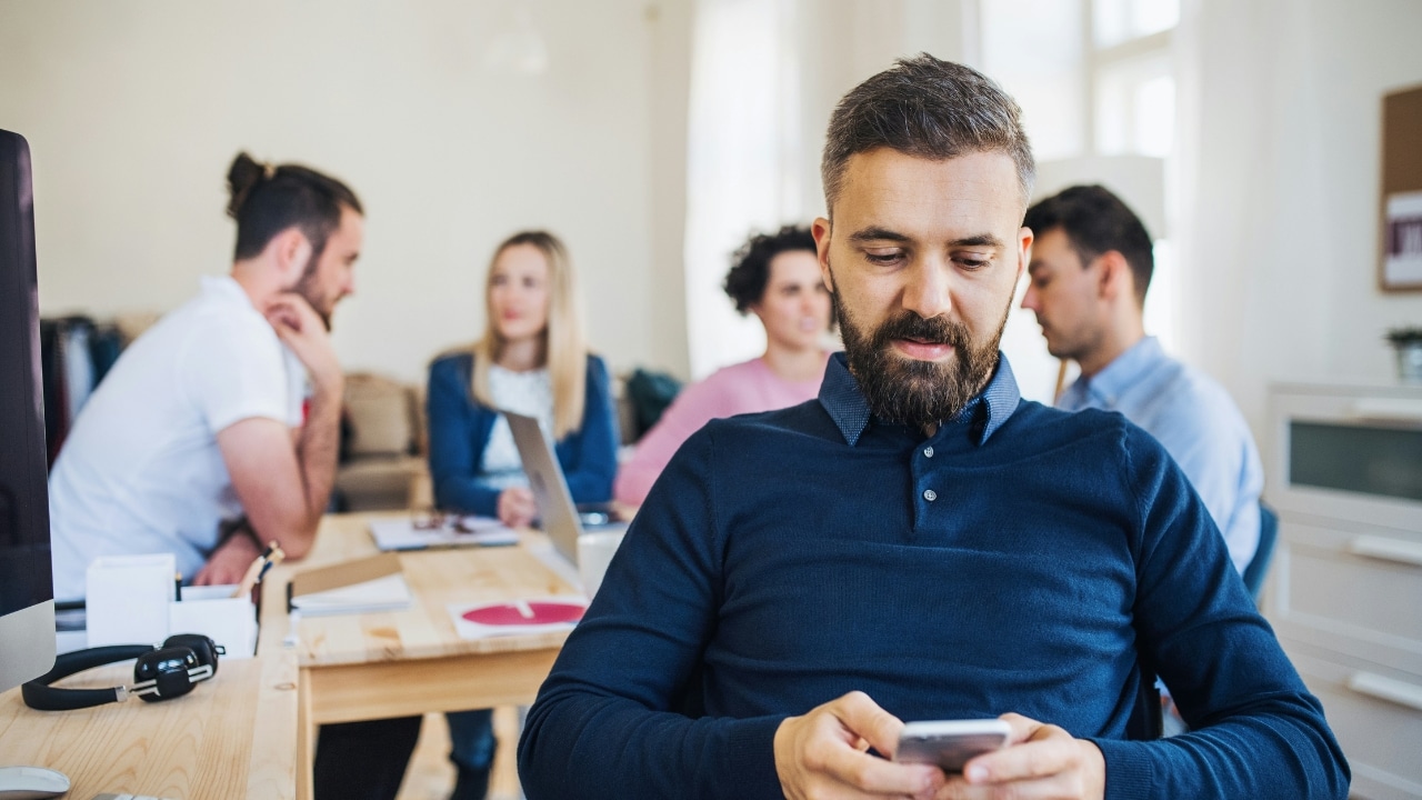 A bearded man in a blue shirt looks at a phone while others sit behind.