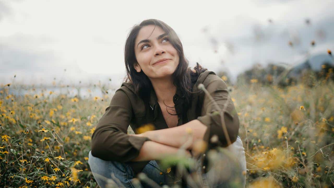 A woman sitting in a wildflower field, looking up with a thoughtful smile.