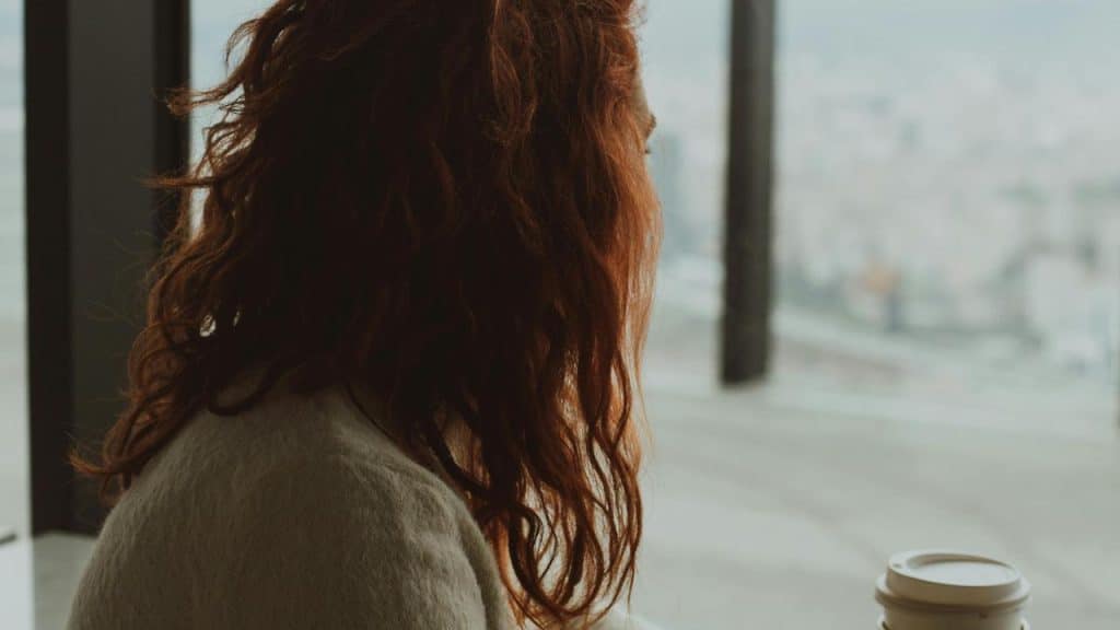 A woman with red hair looking out a window while holding a coffee cup.