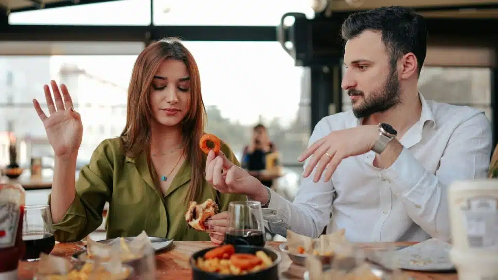 A man offers food to a woman at a restaurant table while she raises her hand.
