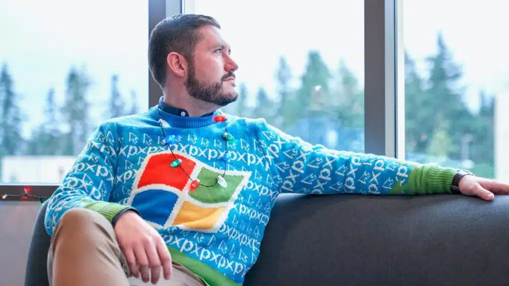 A man with a beard sits on a grey sofa wearing a blue holiday sweater.