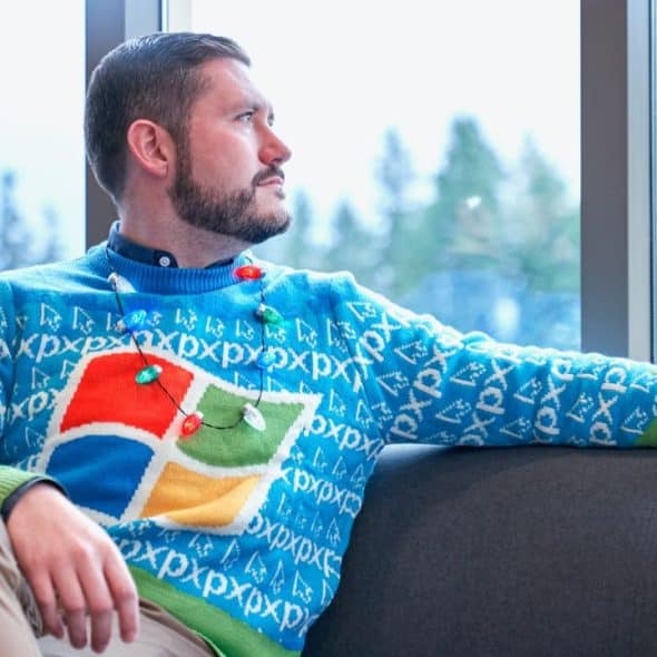 A man with a beard sits on a grey sofa wearing a blue holiday sweater.