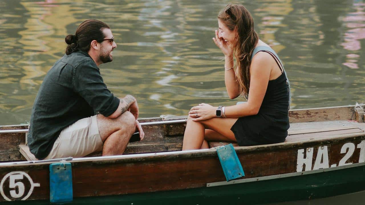 A man with a hair bun and wearing sunglasses smiles as he looks at a laughing woman while they ride on a boat.