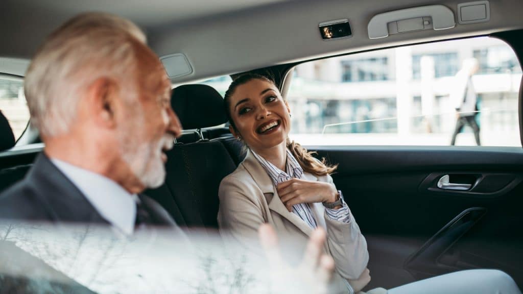 A woman in a light-colored suit smiling and looking at an older man in a car.