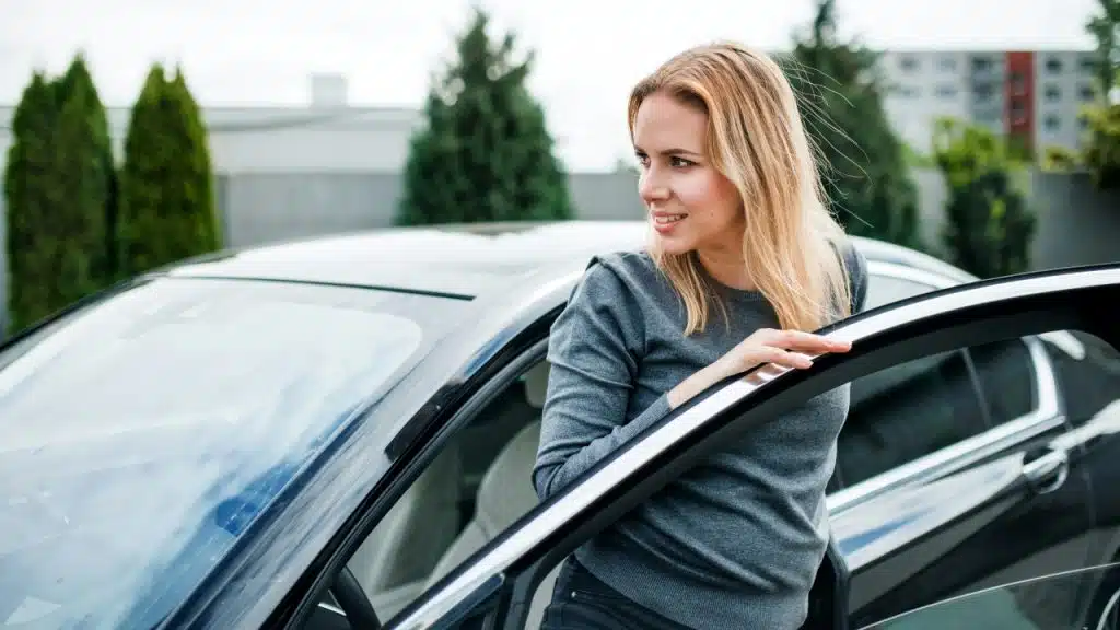 A smiling woman with blonde hair stands by an open car door in a parking lot.