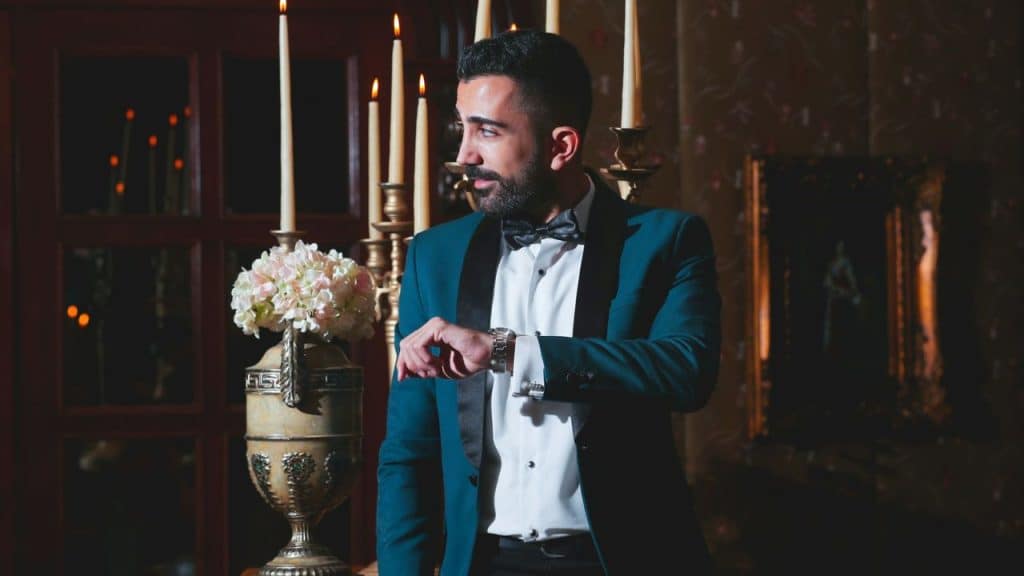 A man in a teal tuxedo checks his watch beside candles and a flower vase.