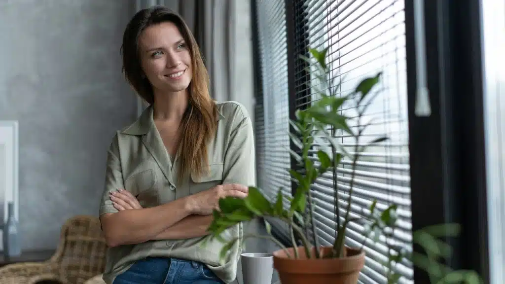 A smiling woman with arms crossed stands by a window with blinds and a plant.