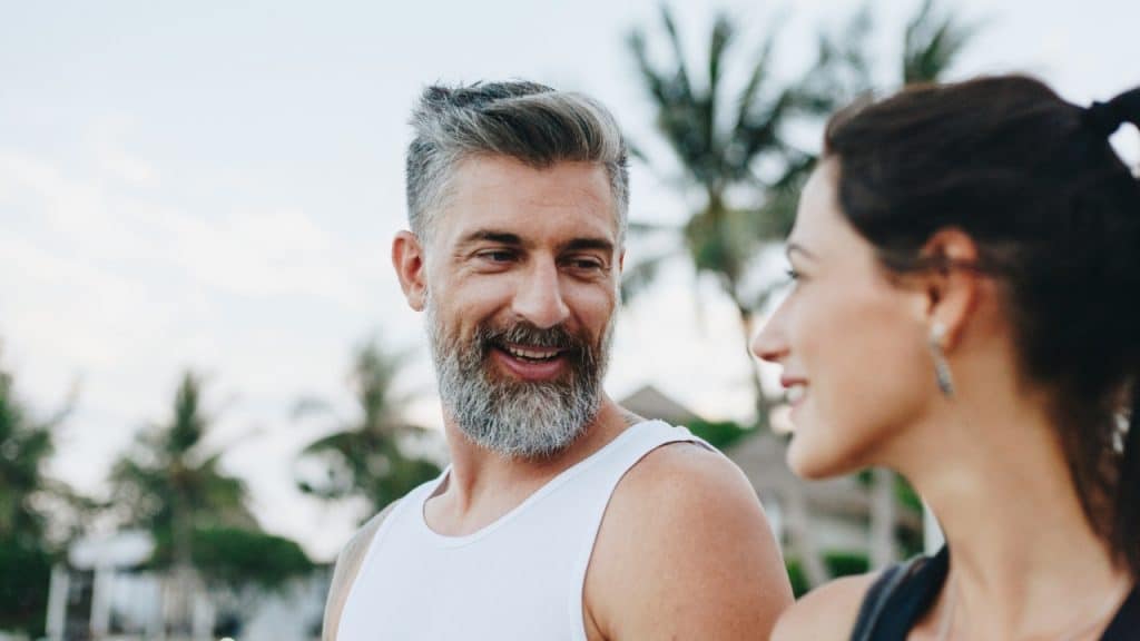 A smiling man with gray hair and a beard looks at a woman near palm trees.