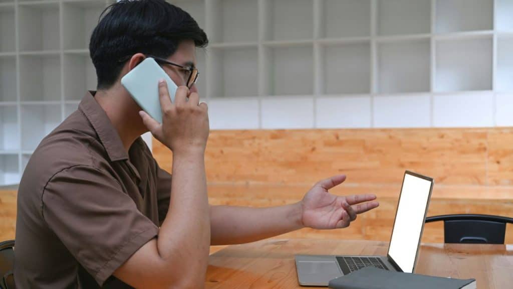 A man talks on a smartphone while gesturing toward an open laptop on a table.