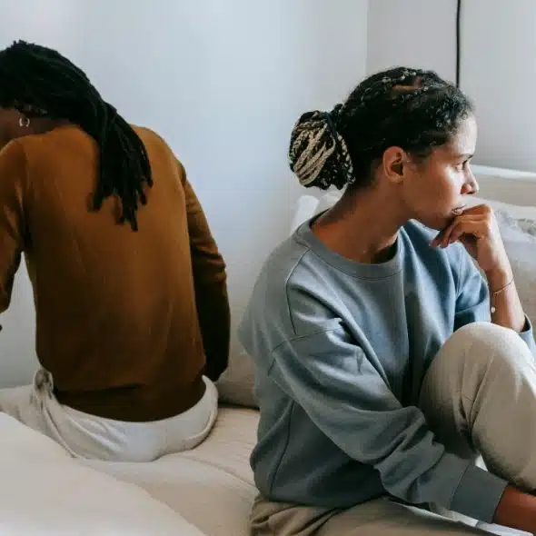 A man and woman sit back-to-back on a bed, looking away in a bright room.