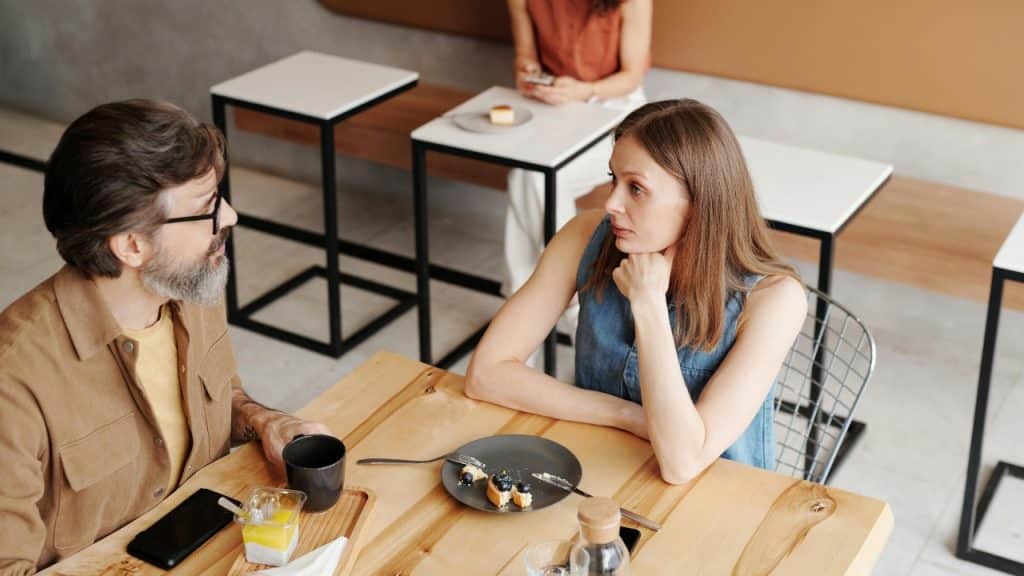 A man and woman sit across from each other at a wooden cafe table talking.