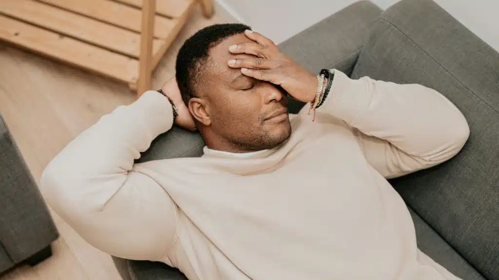 A man in a beige sweatshirt reclining on a gray sofa with his eyes closed.