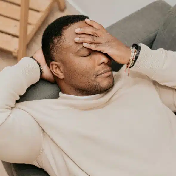 A man in a beige sweatshirt reclining on a gray sofa with his eyes closed.