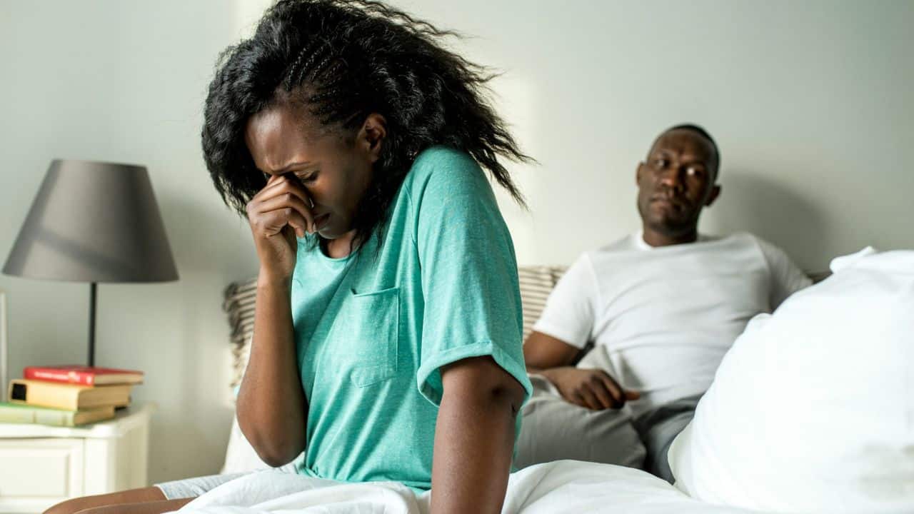 A woman sits on a bed crying into her hand while a man watches behind her.