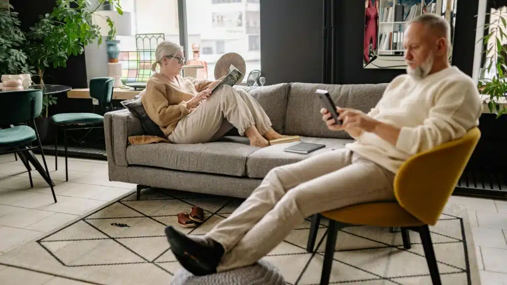 A woman reads a magazine on a sofa while a man uses a mobile phone.