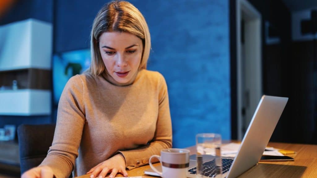 A woman with blonde hair looks down at papers on a desk with a laptop.