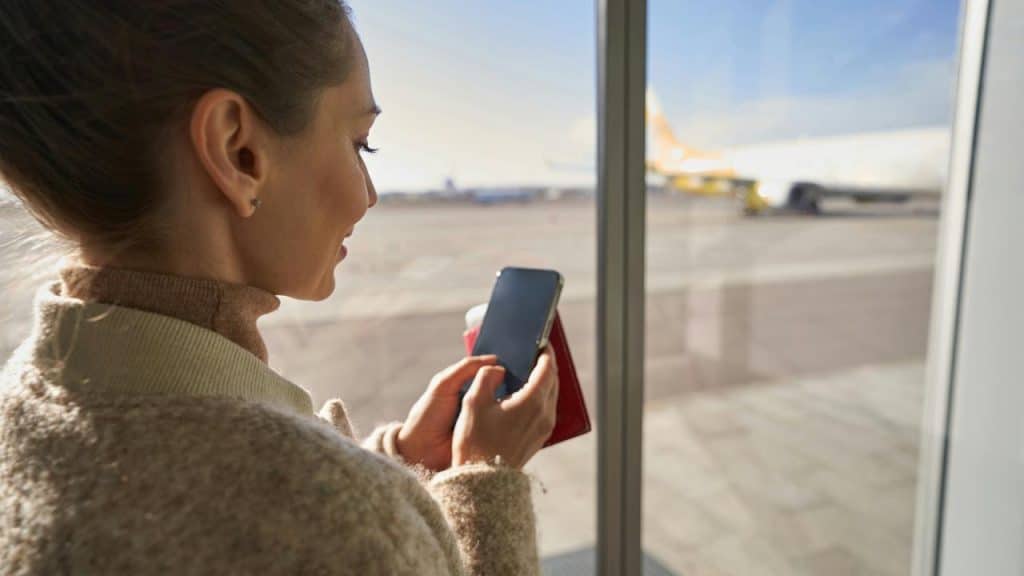 A woman at an airport window holding a phone and passport while looking at a plane outside.