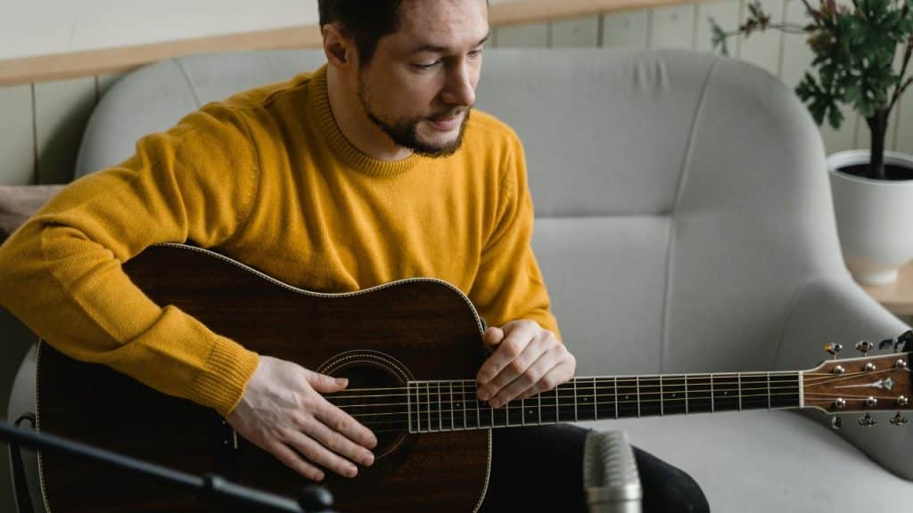A man in a yellow sweater sits on a gray sofa and holds a dark acoustic guitar.