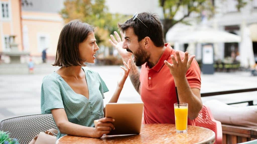 A man and woman sit at an outdoor table gesturing and speaking intensely to each other.