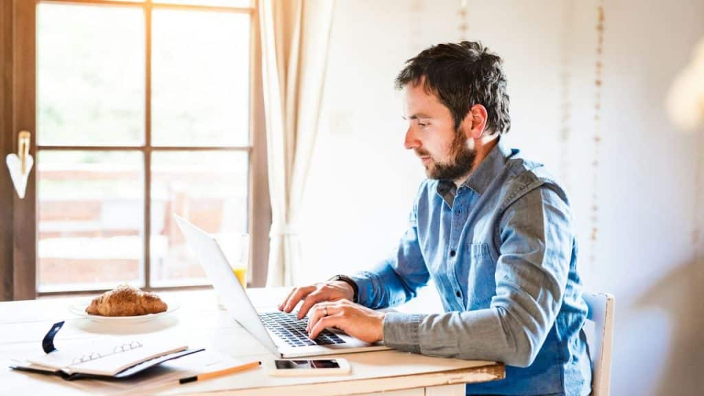 A man sits at a white table typing on a laptop near a window.