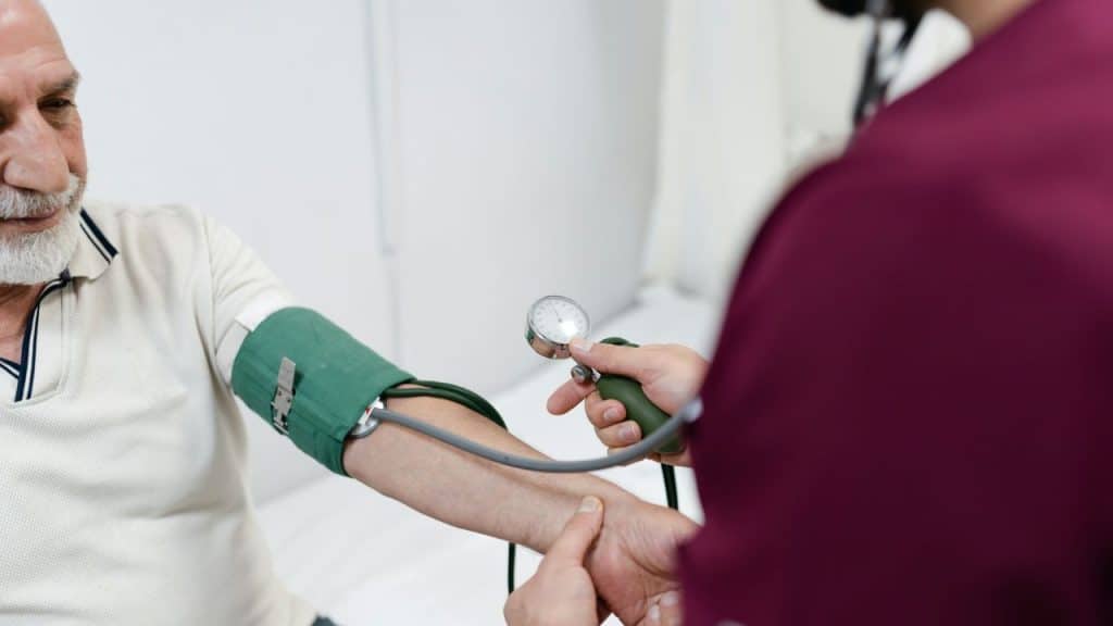 A person in a purple shirt uses a manual blood pressure monitor on an older man's arm.