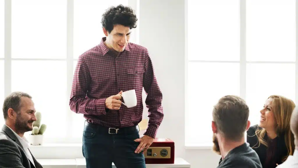 A man in a plaid shirt holds a white mug while talking to a seated group.