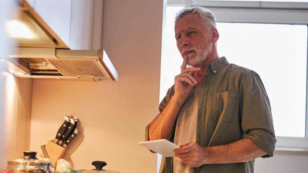 A middle-aged man with a gray beard holds a tablet, touching his chin while thinking.