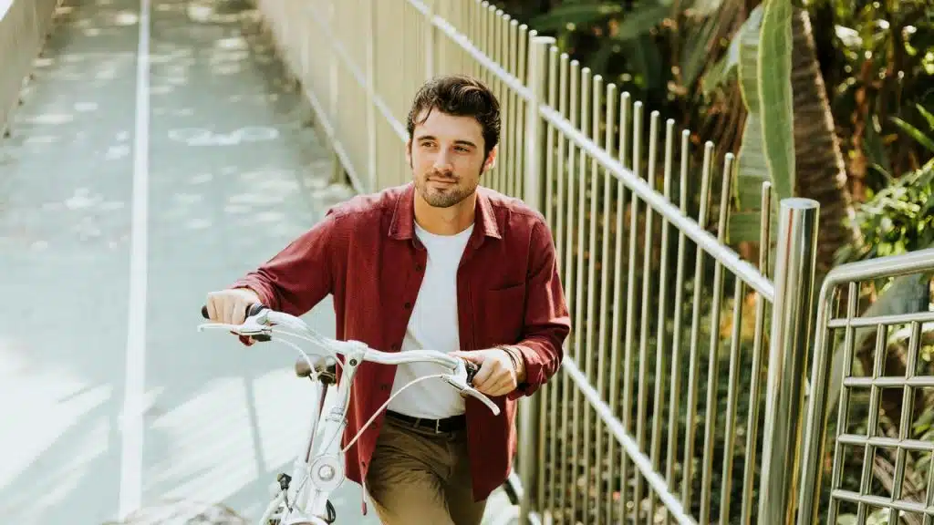 A man walking a bicycle along a sunlit path beside a metal fence.