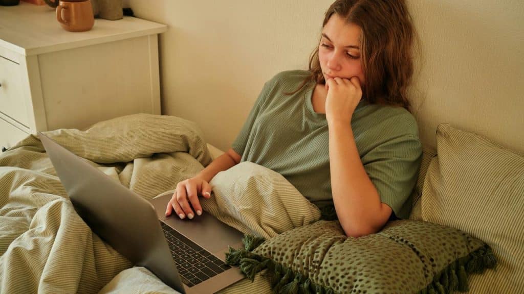 A woman lying in bed using a laptop, resting her chin on her hand.