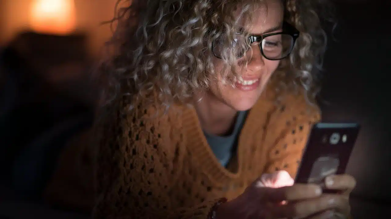 A woman with curly hair and glasses smiling at her smartphone in low light.