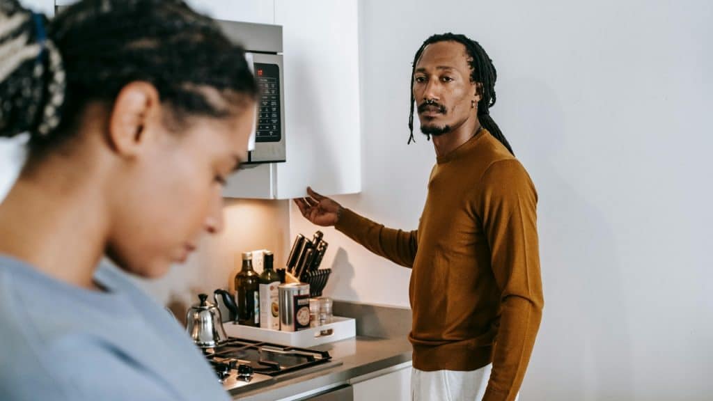 A man opens a kitchen cabinet while a woman stands in the foreground, looking down.