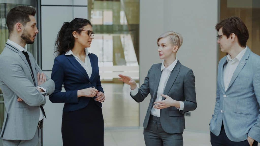 Four professionals in business attire stand in an office lobby during a work discussion.