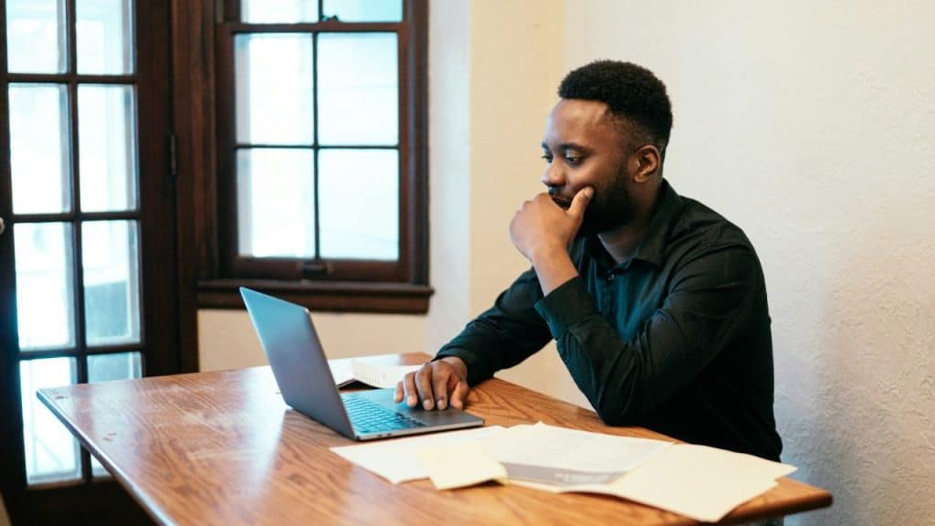 A man in a black shirt sits at a wooden table looking at a laptop.