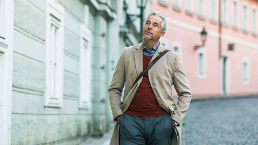 A man with gray hair in a beige coat looks upward while walking.