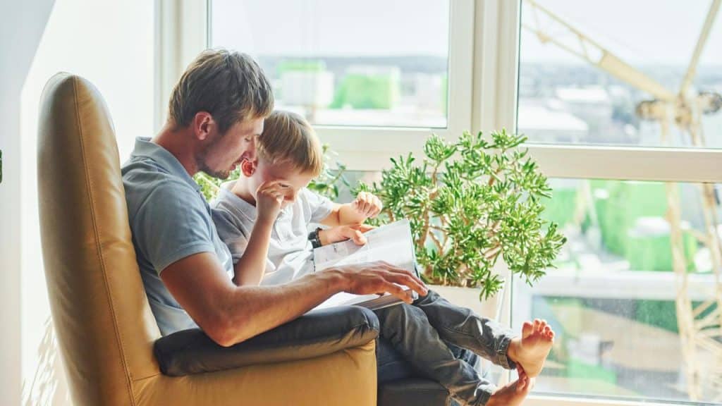 A man sitting in a chair reading a book with a young boy by the window.