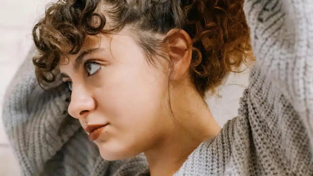 A curly-haired person in a sweater tying their hair up, shown in side profile.