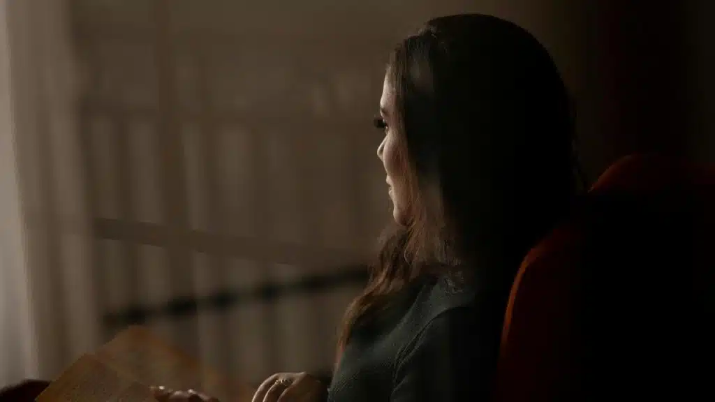 A woman sitting by a window holding a book and looking outside in soft light.
