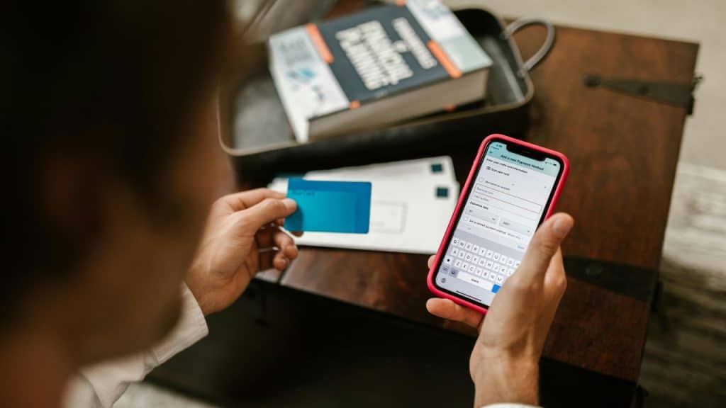 A man holds a blue credit card while typing payment details into his phone.