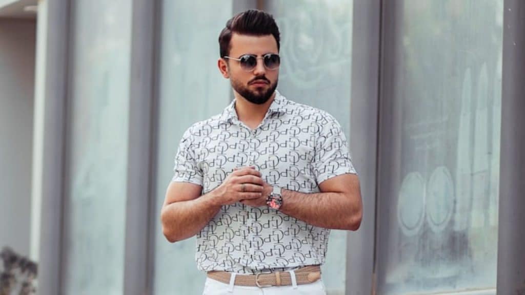 A man in a patterned shirt and sunglasses poses with a watch on his wrist.