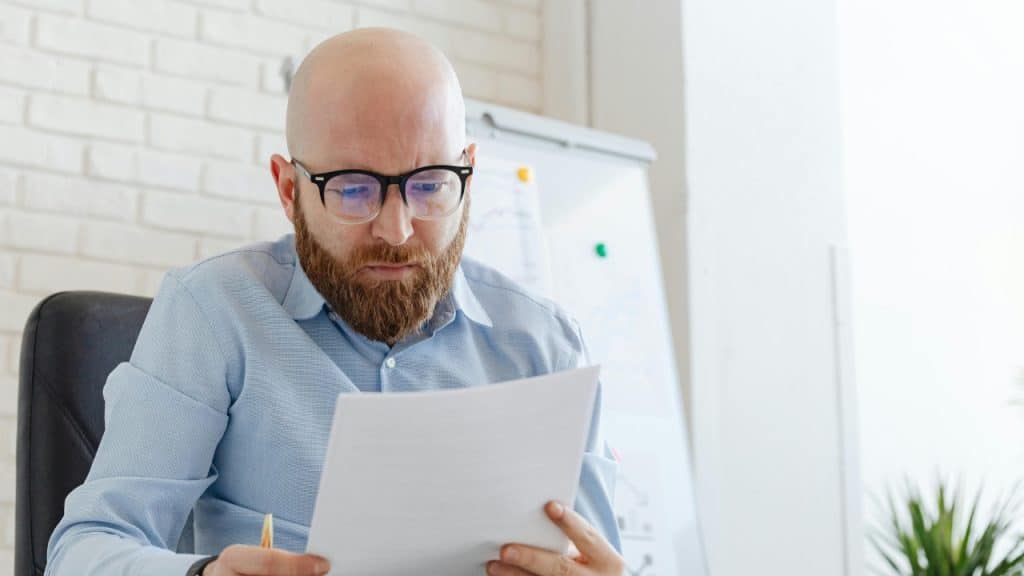 A man with a beard and glasses sits while intently reading a document.