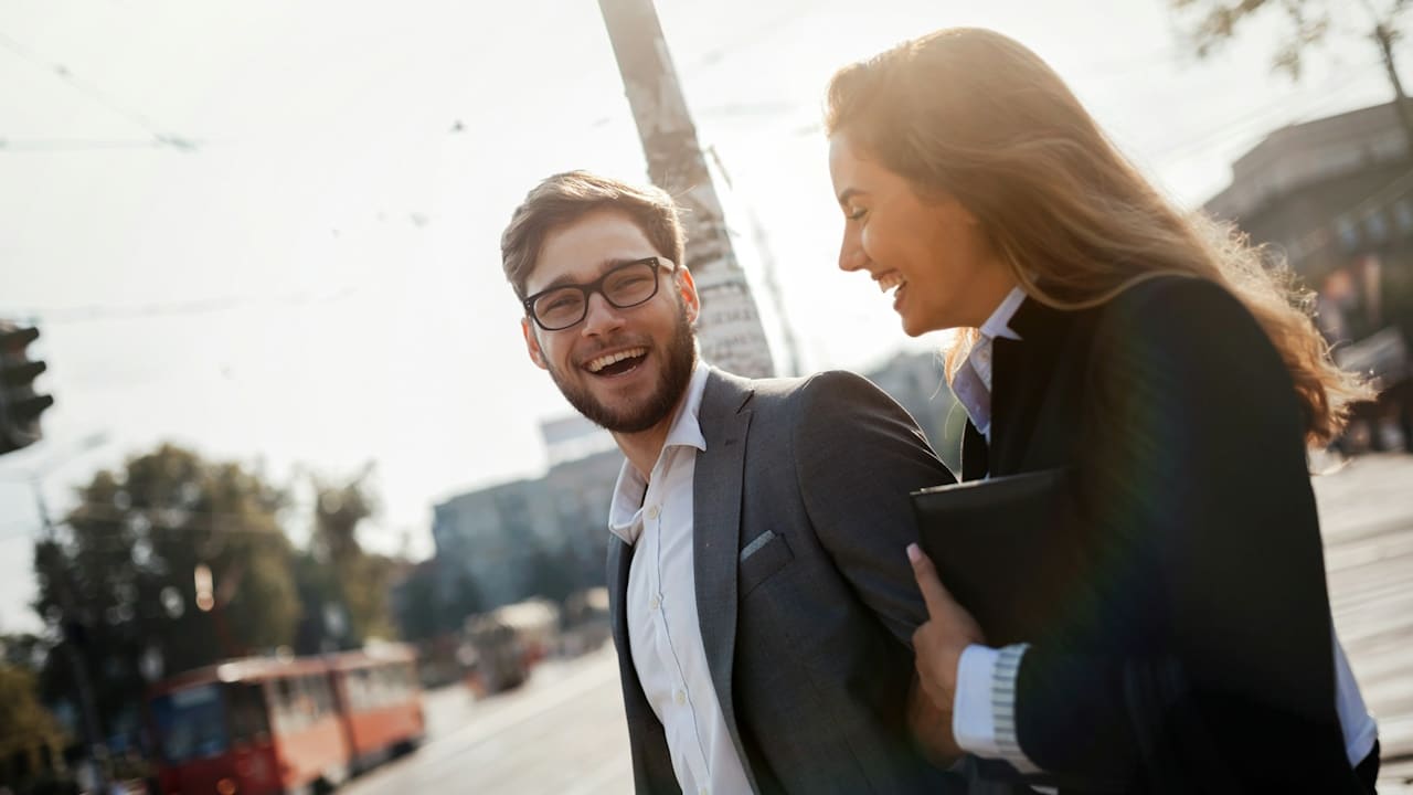 A bearded, bespectacled man wearing a suit smiling while walking on a street with a smiling woman who holds a file by his side. 