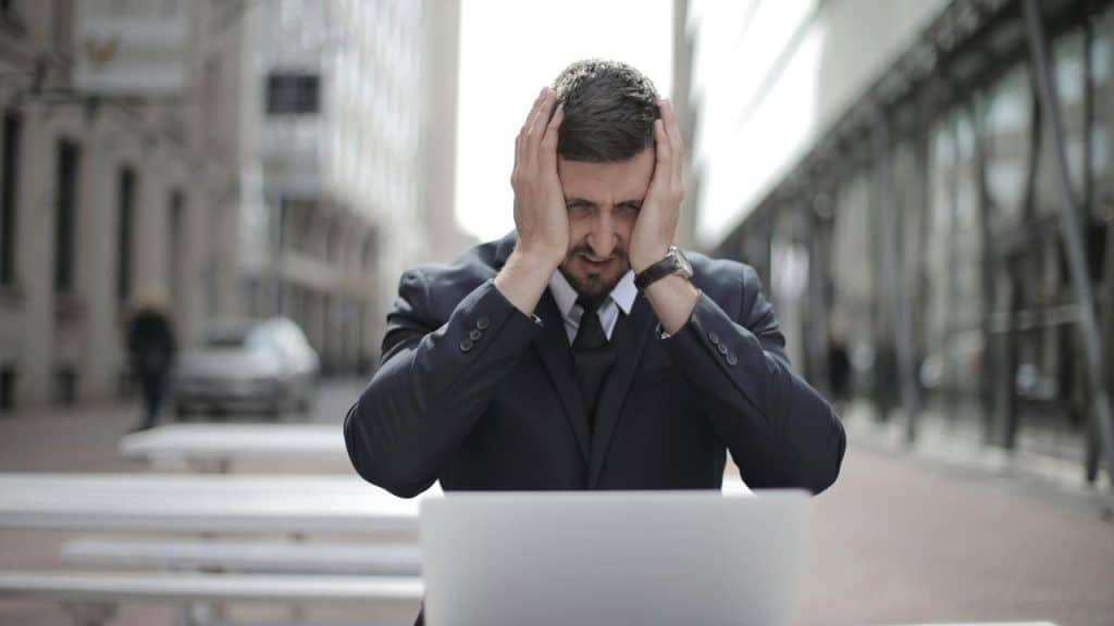 A stressed businessman holding his head while sitting in front of a laptop outdoors.