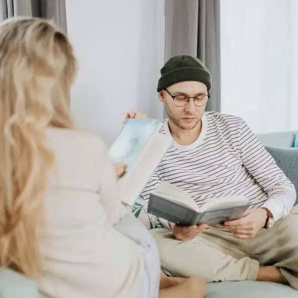 A man and a woman sitting on a couch reading books together in a bright living room.