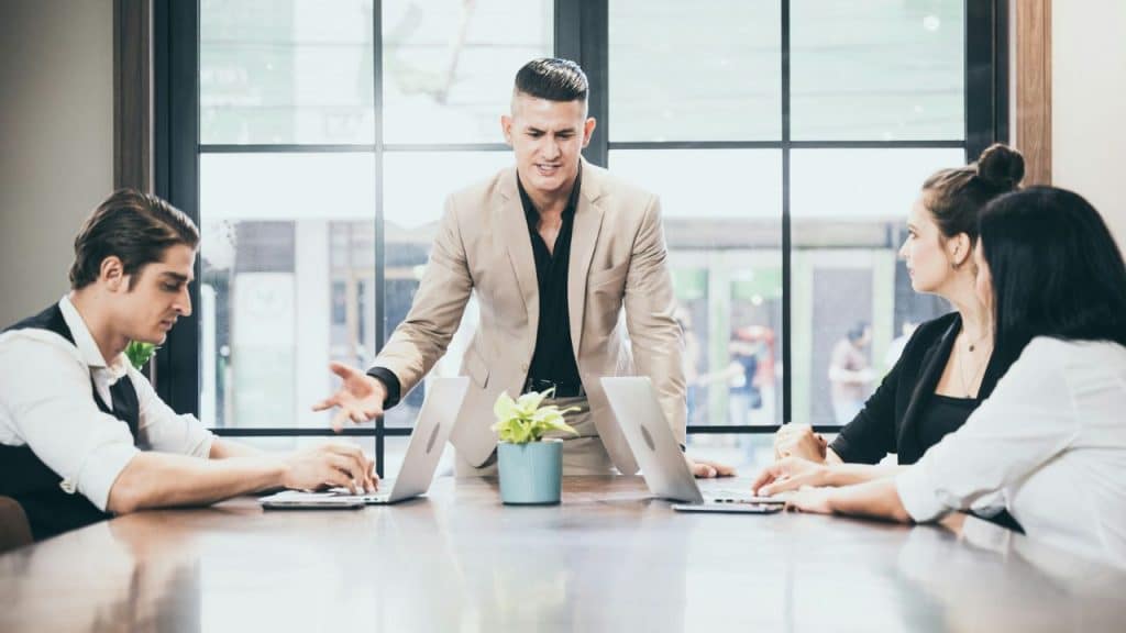 A man in a beige suit stands while talking to three people at a table.