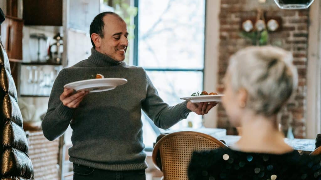 A man in a gray turtleneck holds two plates of food while facing a woman.