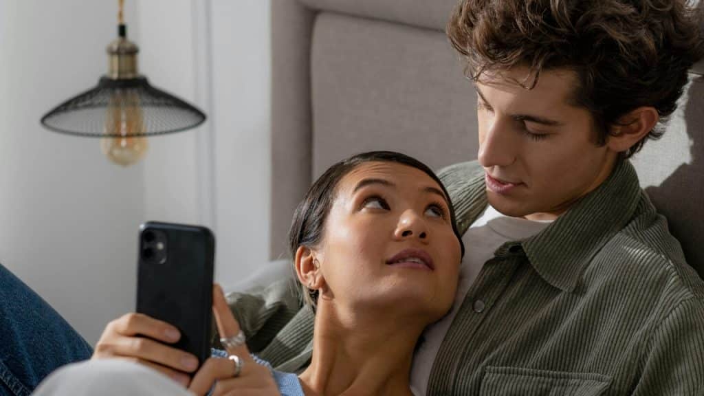 A woman holds a smartphone while lying down, looking at a man nearby.
