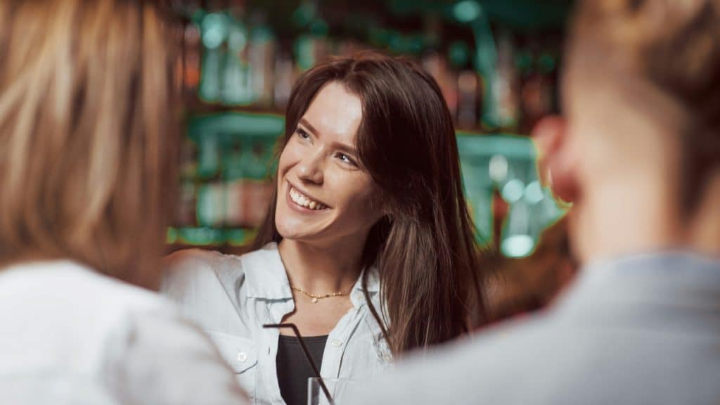 A woman with long brown hair smiles while talking to people in a dimly lit room.