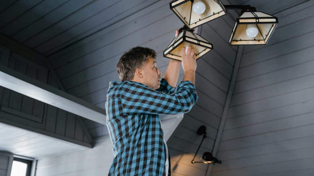 A man in a plaid shirt stands on a ladder, changing a lightbulb overhead.